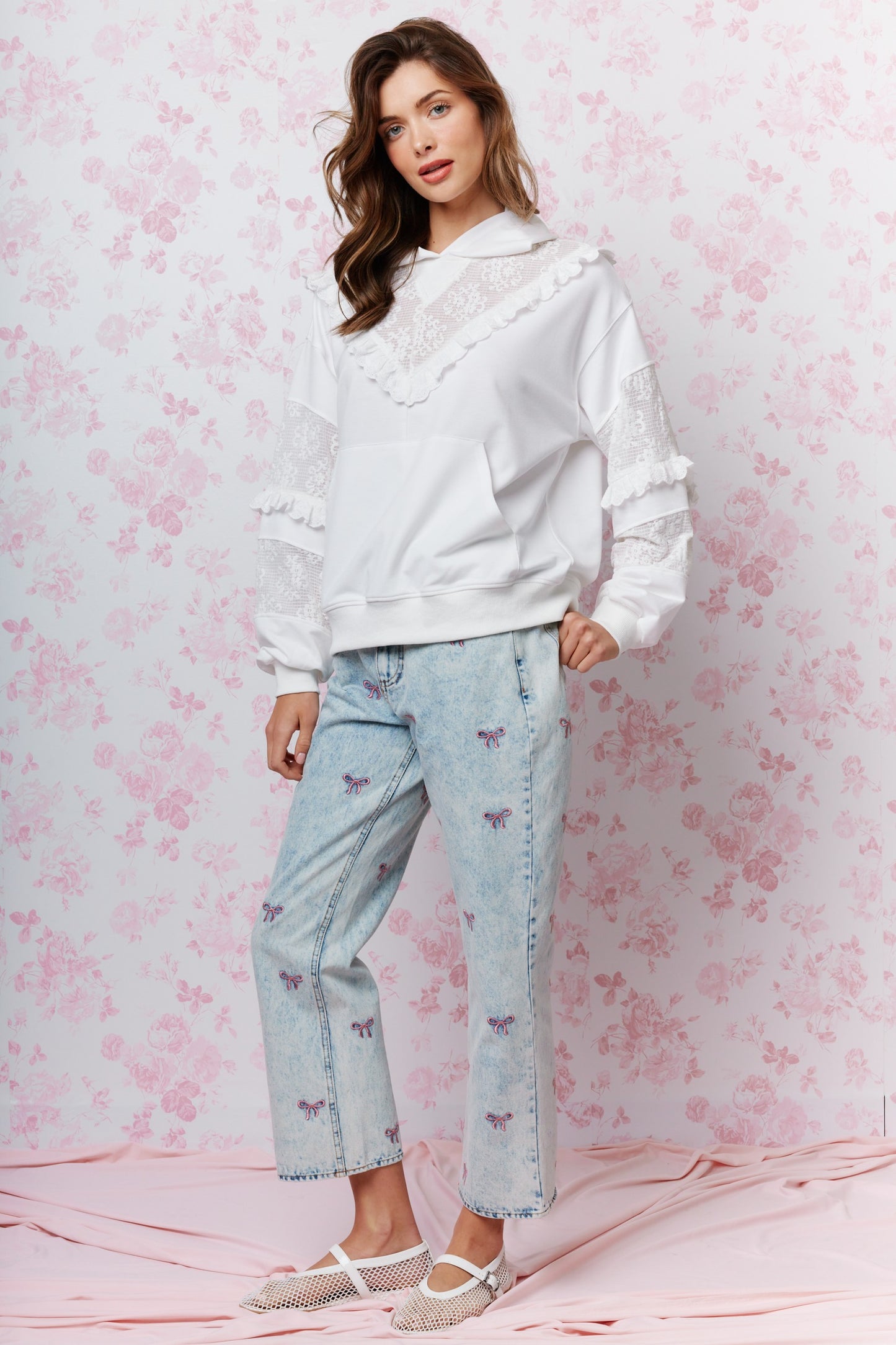 A woman models a white lace-trimmed hoodie and light-wash blue jeans with butterfly embroidery, standing against a pink floral wallpaper background.