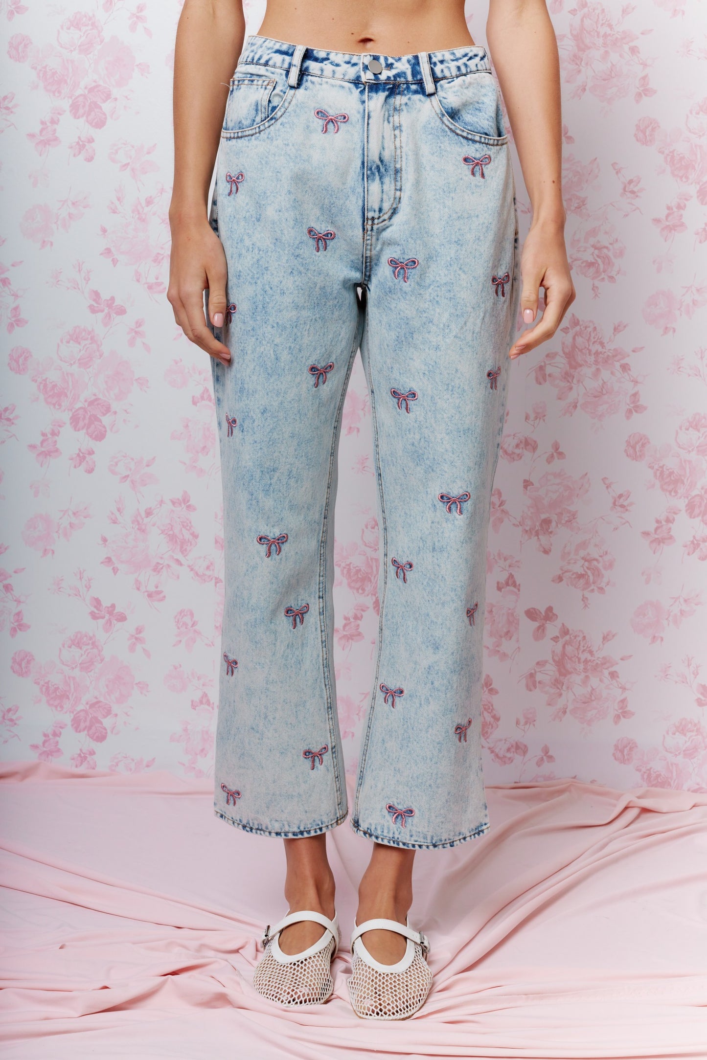 A model is wearing light-wash blue cropped jeans with an all-over embroidered butterfly pattern, paired with white strappy flat shoes, standing against a pink floral backdrop.