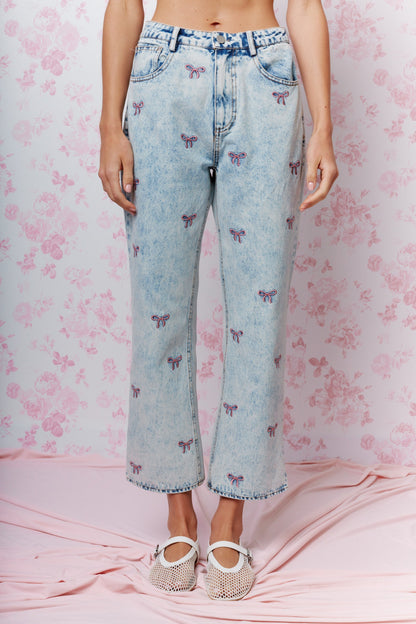 A model is wearing light-wash blue cropped jeans with an all-over embroidered butterfly pattern, paired with white strappy flat shoes, standing against a pink floral backdrop.