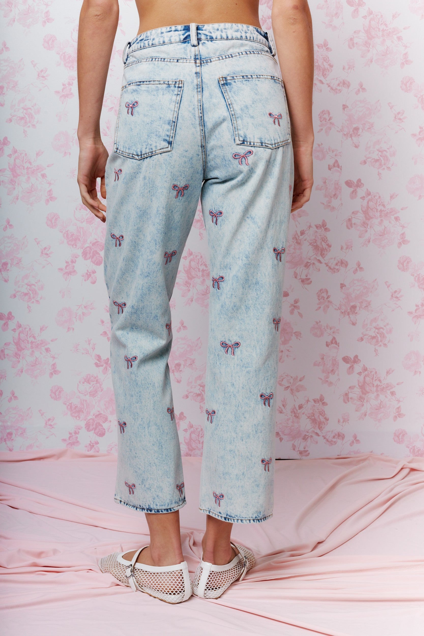 Back view of a person wearing light blue cropped denim jeans with a small purple butterfly print, standing against a pink floral backdrop.