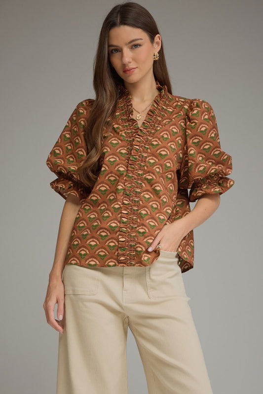 A woman modeling a brown short-sleeve blouse with a green and beige scalloped fan pattern, paired with cream-colored trousers, against a solid gray studio background.