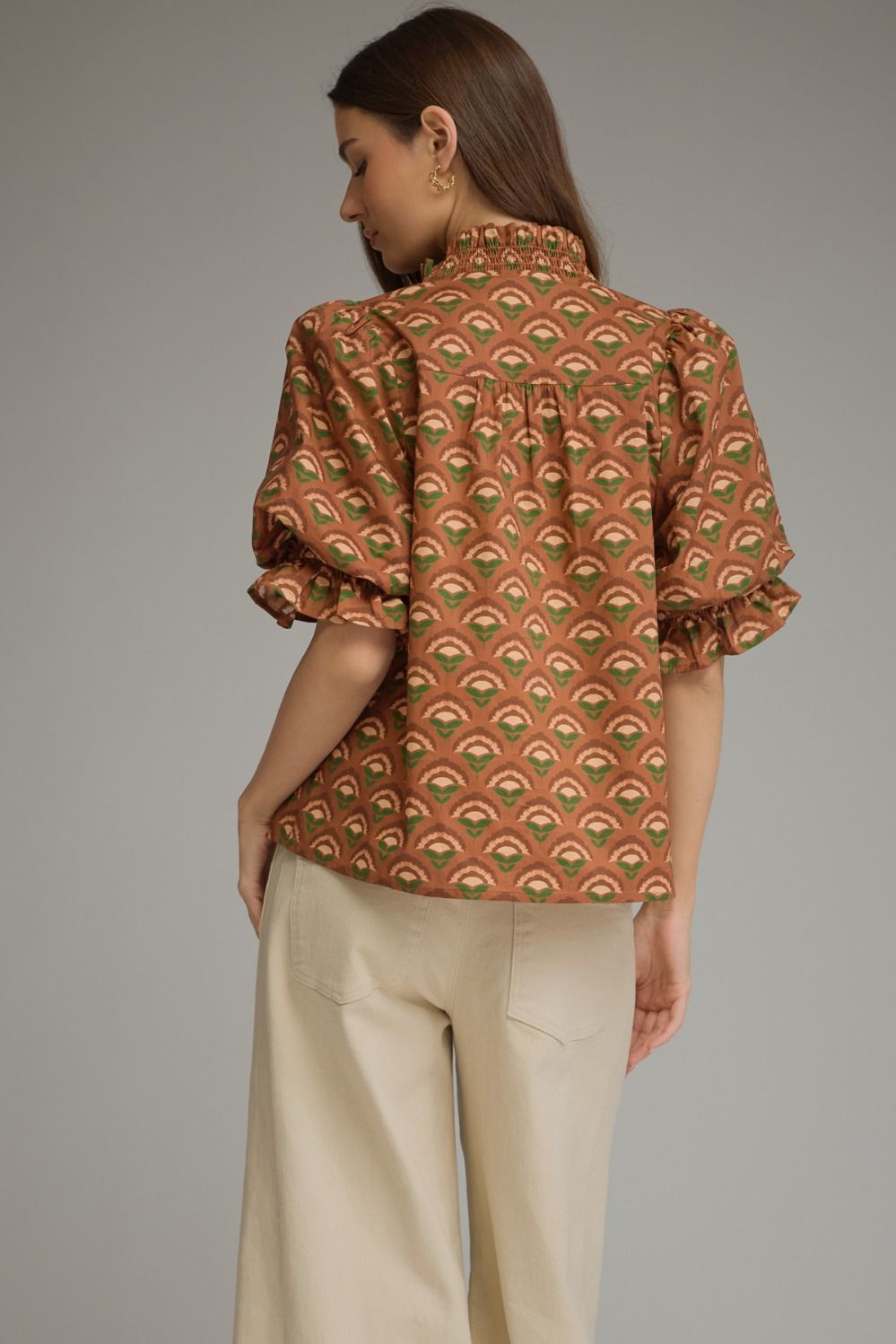 A woman shown from the back wearing a rust-orange short-sleeve blouse with a green and beige repeating fan pattern, paired with light beige trousers, against a solid gray studio background.