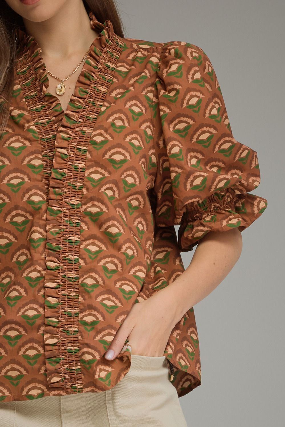 A close-up of a woman wearing a brown and green patterned blouse with ruffled trim and puff sleeves, paired with light beige pants against a plain gray background.