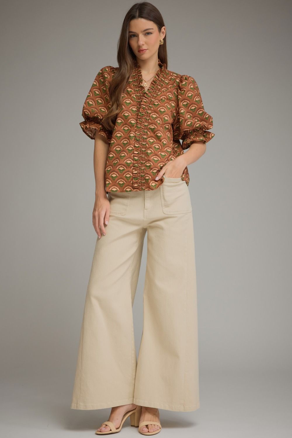 A woman modeling a brown and gold patterned puff-sleeve blouse paired with wide-leg cream pants, standing against a neutral gray studio background.