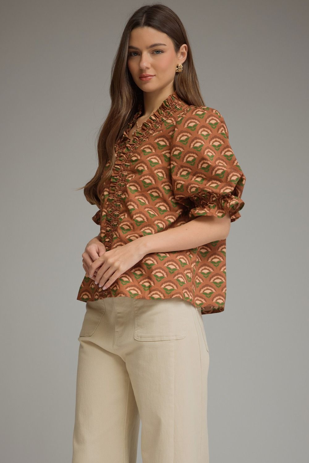 A woman modeling a brown short-sleeve blouse with a green and beige repeating leaf pattern, paired with cream-colored trousers, against a solid gray studio background.