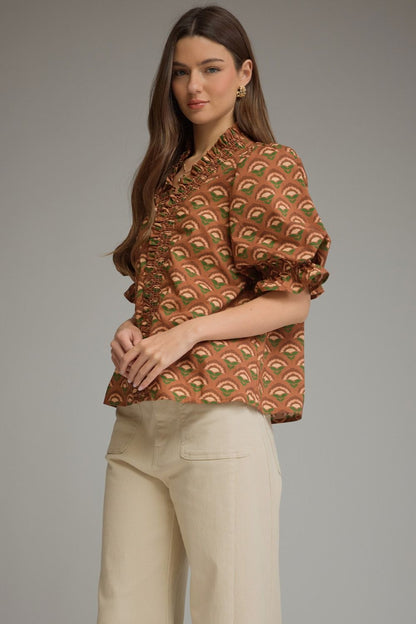 A woman modeling a brown short-sleeve blouse with a green and beige repeating leaf pattern, paired with cream-colored trousers, against a solid gray studio background.