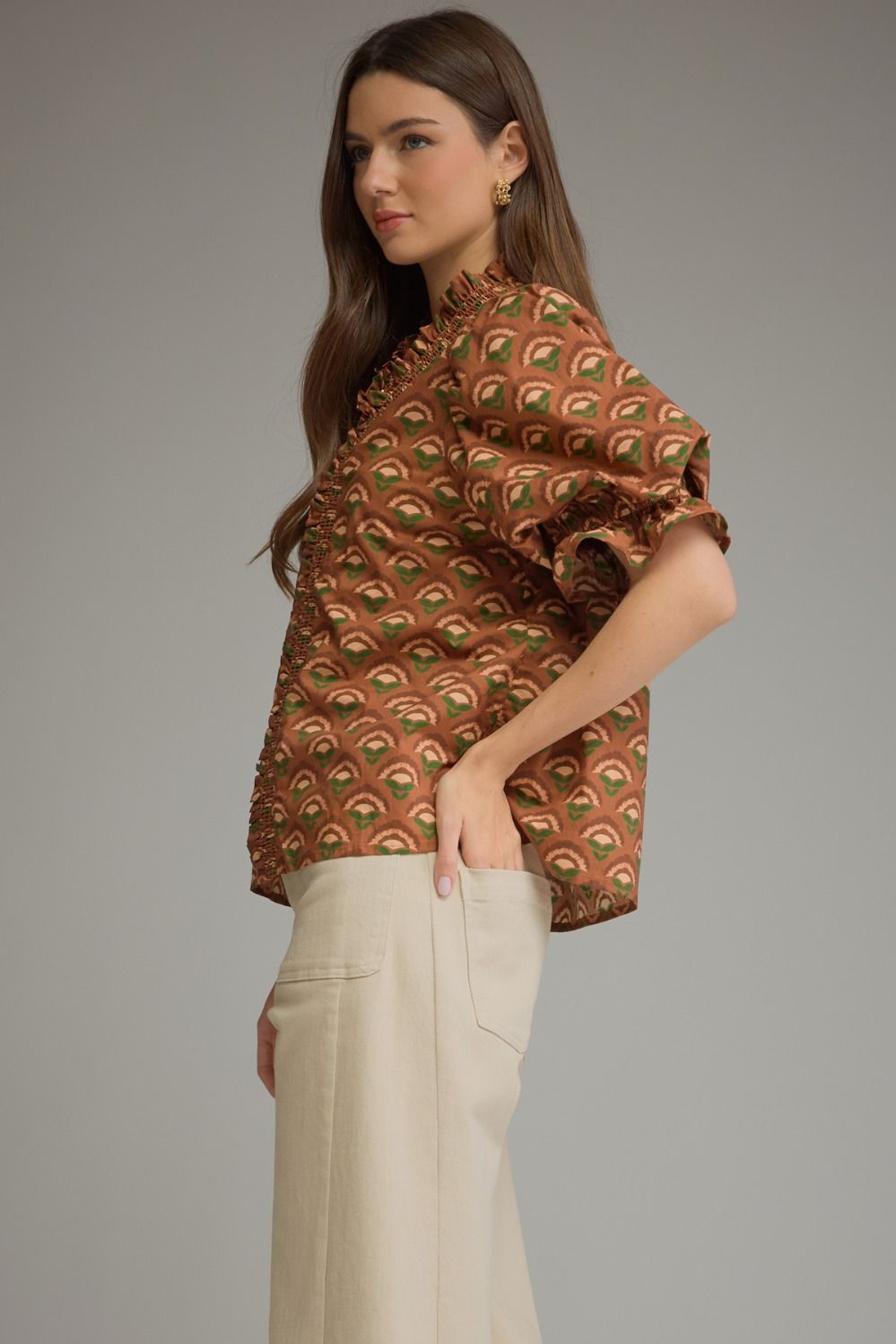 A woman modeling a brown short-sleeve blouse with a green and beige peacock feather pattern, paired with cream-colored trousers, against a solid gray studio background.
