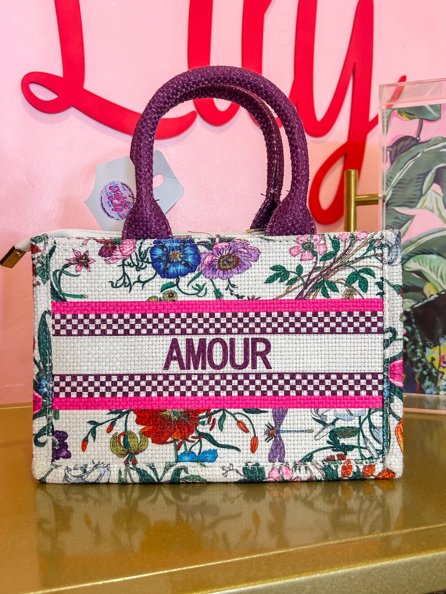 Floral embroidered tote bag with purple handles and 'Amour' text, displayed on gold shelf.