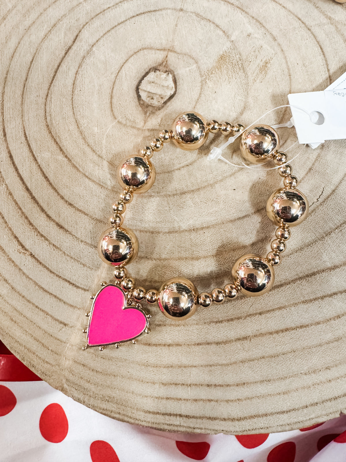 Gold beaded bracelet with a pink heart charm on a wooden background, jewelry accessory