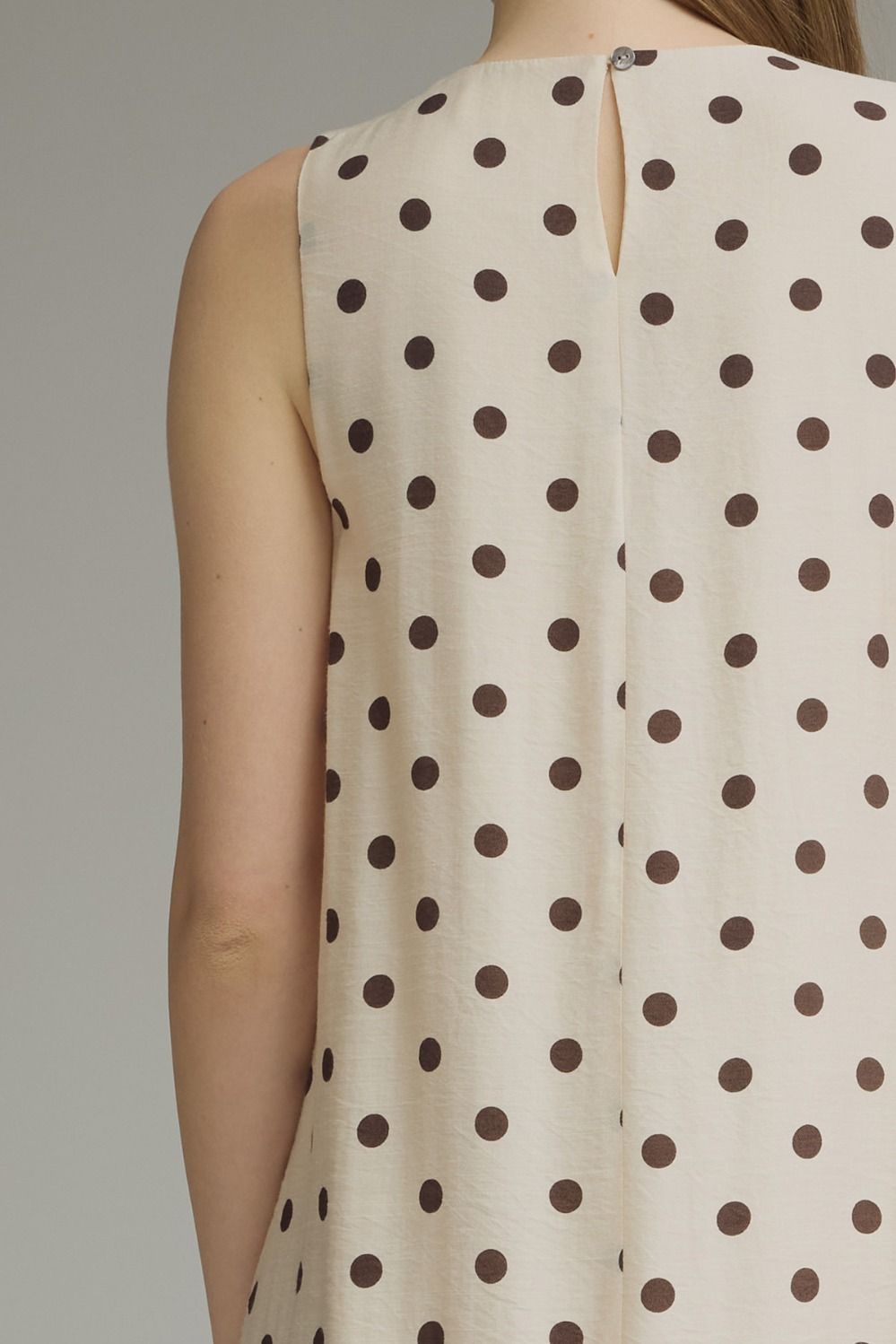 Close-up back view of a sleeveless top or dress with an off-white base and dark brown polka dots, shown on a model against a plain gray studio background.