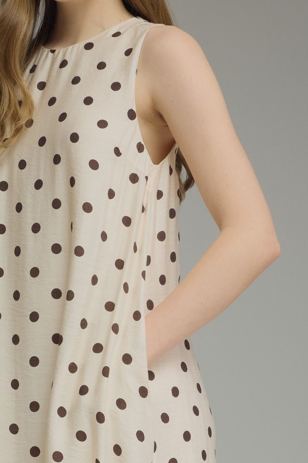 Close-up of a sleeveless cream-colored dress with dark brown polka dots, worn by a model against a solid gray background.