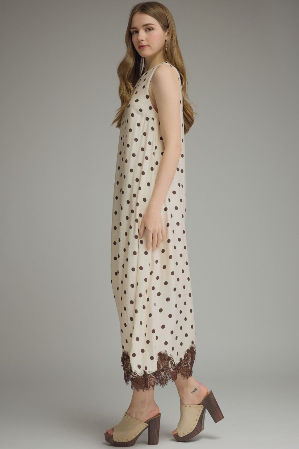 A woman modeling a sleeveless cream maxi dress with brown polka dots and a brown lace hem, paired with beige and brown block-heel mule sandals, against a neutral gray studio background.