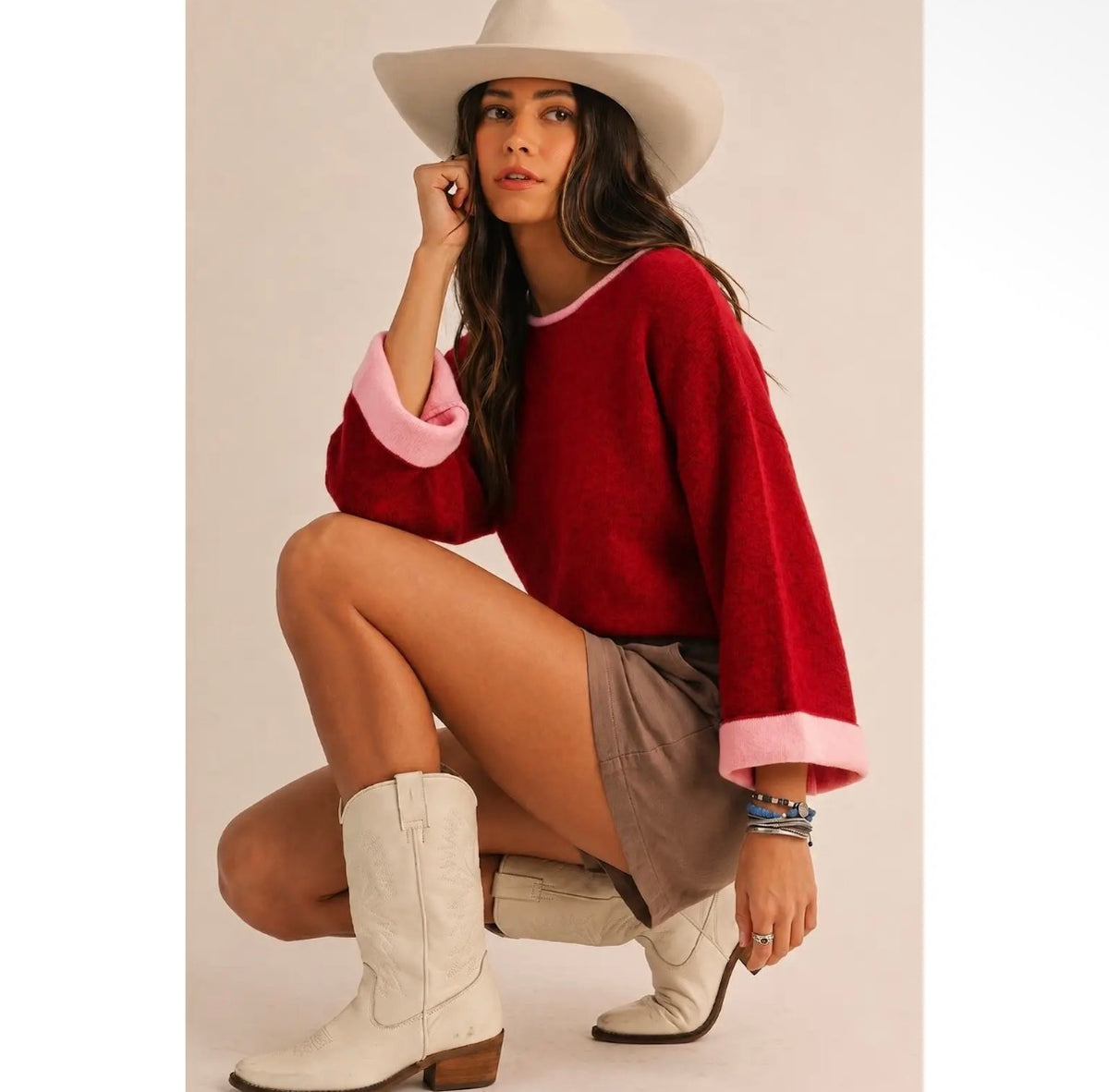 Model wearing cherry red relaxed-fit women's knit sweater with bubblegum pink fold-over contrast cuffs and pink neckline, styled with a mini skirt and western boots.