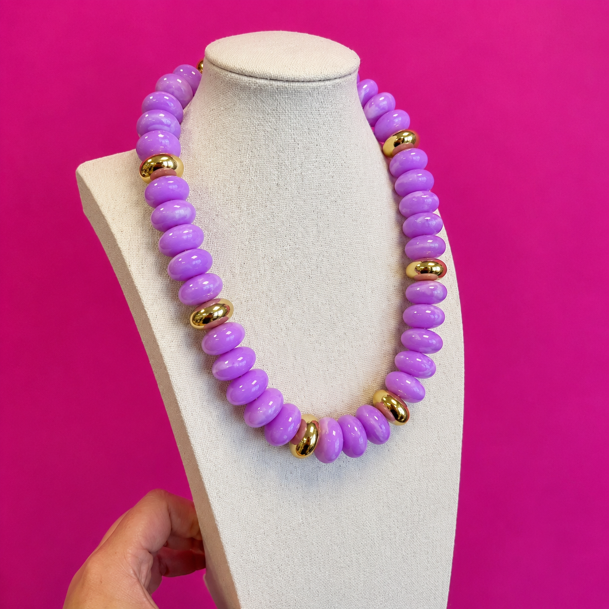 A chunky statement necklace made of glossy lavender-purple beads with small polished gold oval accents, displayed on a cream jewelry bust against a bright pink background. A hand is visible at the bottom left holding the display stand.