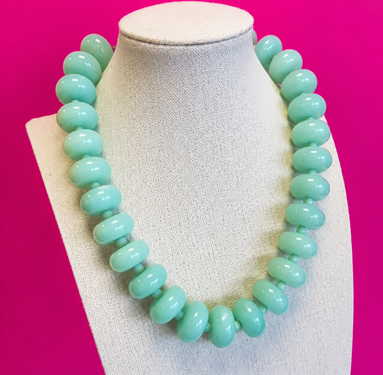 A chunky beaded necklace in a light mint green color, displayed on a textured off-white fabric jewelry bust against a solid hot pink background.