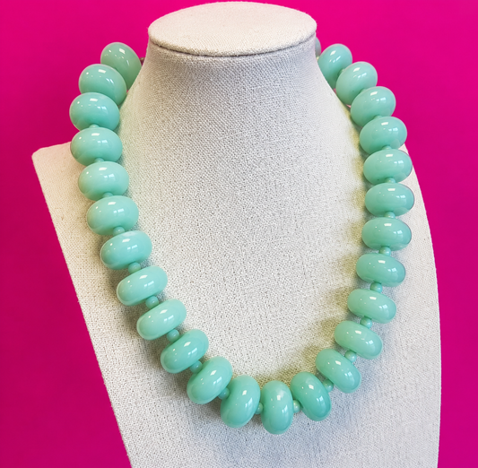 A chunky beaded necklace in a light mint green color, displayed on a textured off-white fabric jewelry bust against a solid hot pink background.