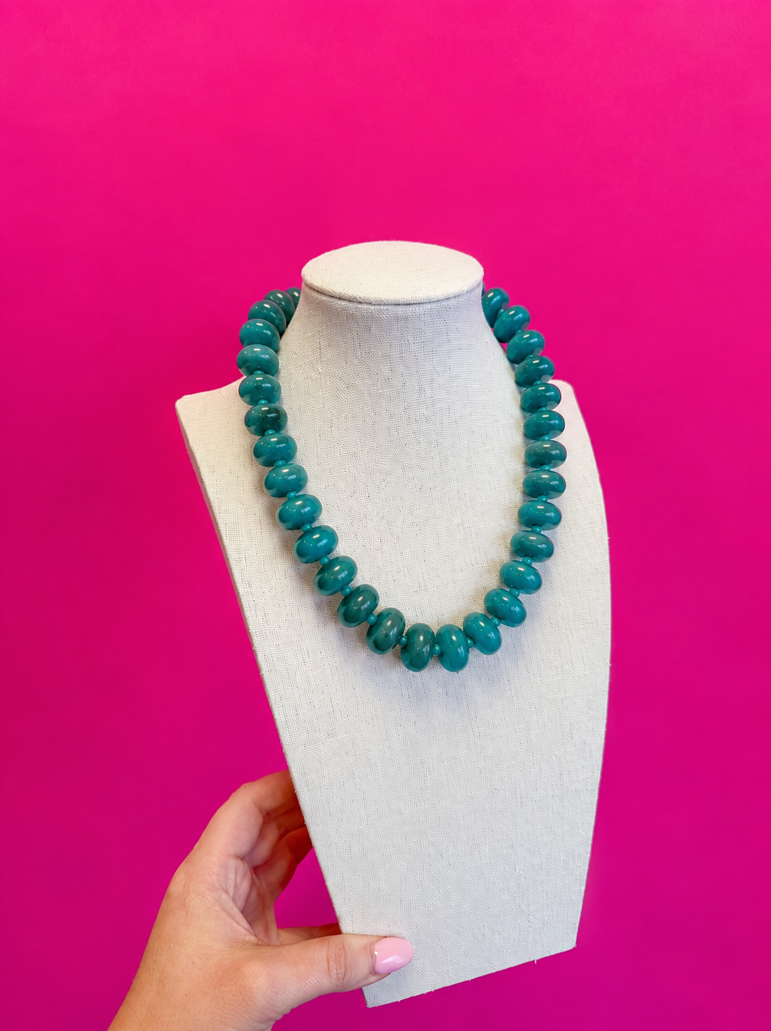 A turquoise beaded necklace displayed on a cream-colored jewelry bust, held at the bottom by a hand against a solid hot pink background.