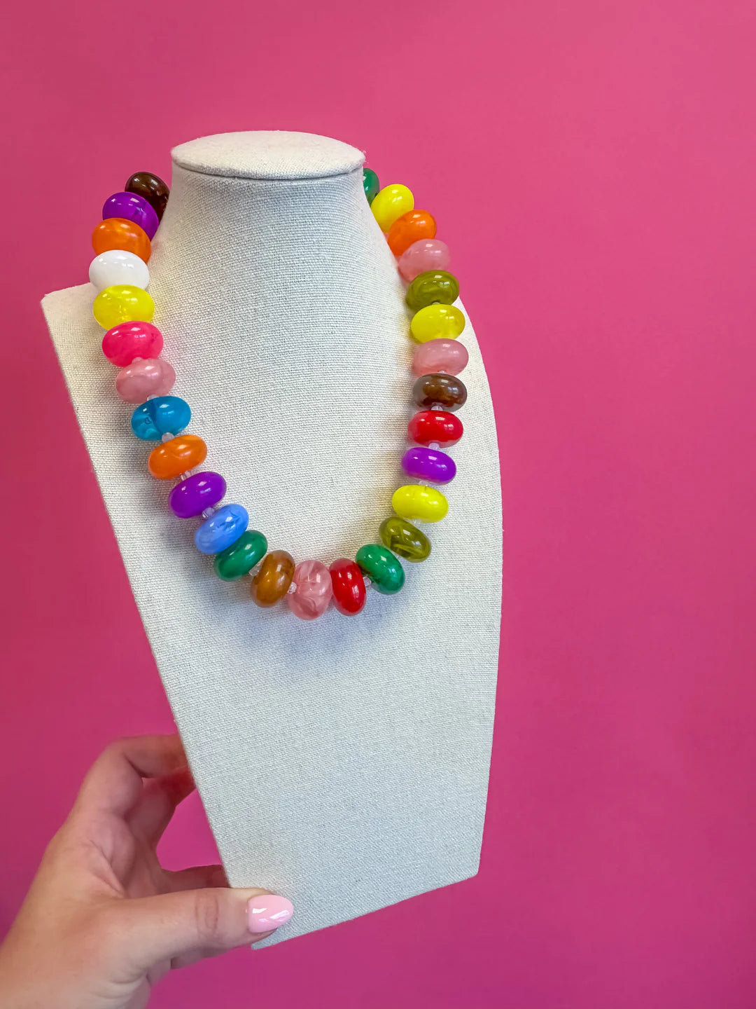 A multicolor beaded necklace made of glossy, translucent jelly bean-shaped beads, displayed on a light gray fabric jewelry bust held by a hand against a solid pink background.