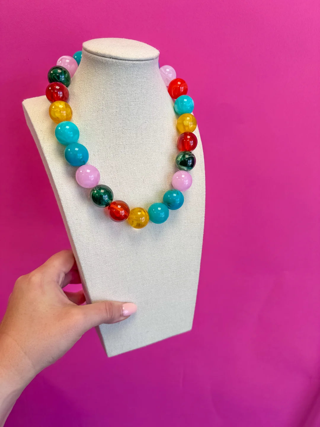 A colorful beaded necklace displayed on a cream jewelry bust, held by a hand against a bright pink background.