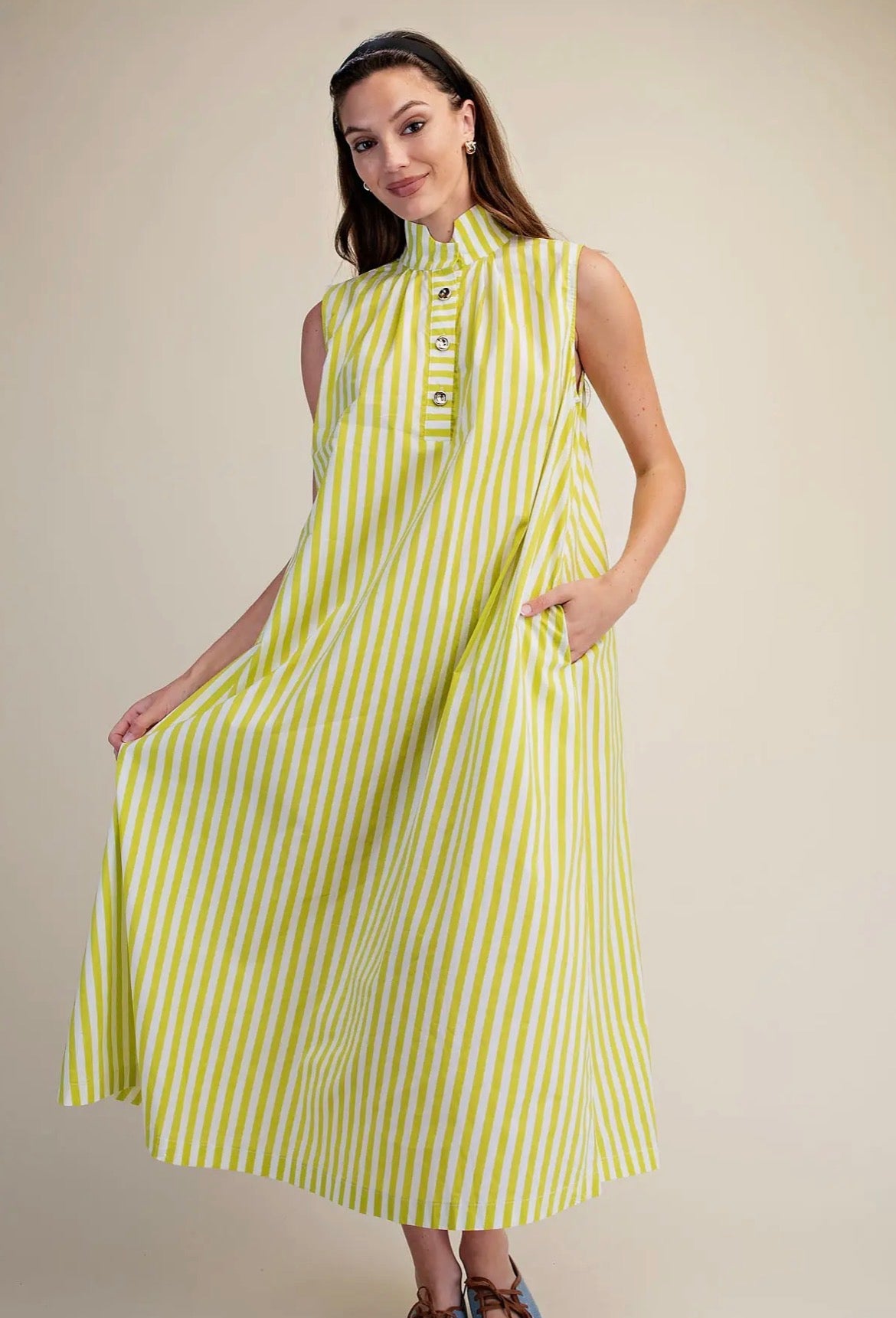 A woman modeling a sleeveless midi dress with vertical lime green and white stripes, featuring a mock neck and decorative buttons at the collar, set against a plain beige studio background.