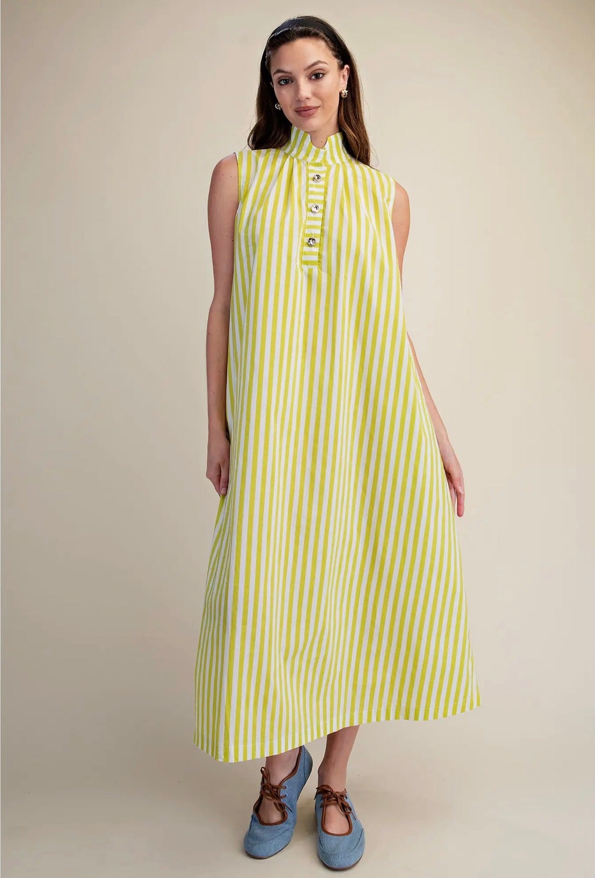 A woman modeling a sleeveless midi dress with vertical lime green and white stripes, featuring a mock neck and decorative button details at the collar, paired with blue shoes against a solid beige background.