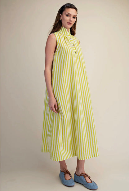 A woman modeling a sleeveless midi dress with vertical lime green and white stripes, featuring a high mock neck and buttoned front, paired with blue Mary Jane shoes against a solid beige studio background.