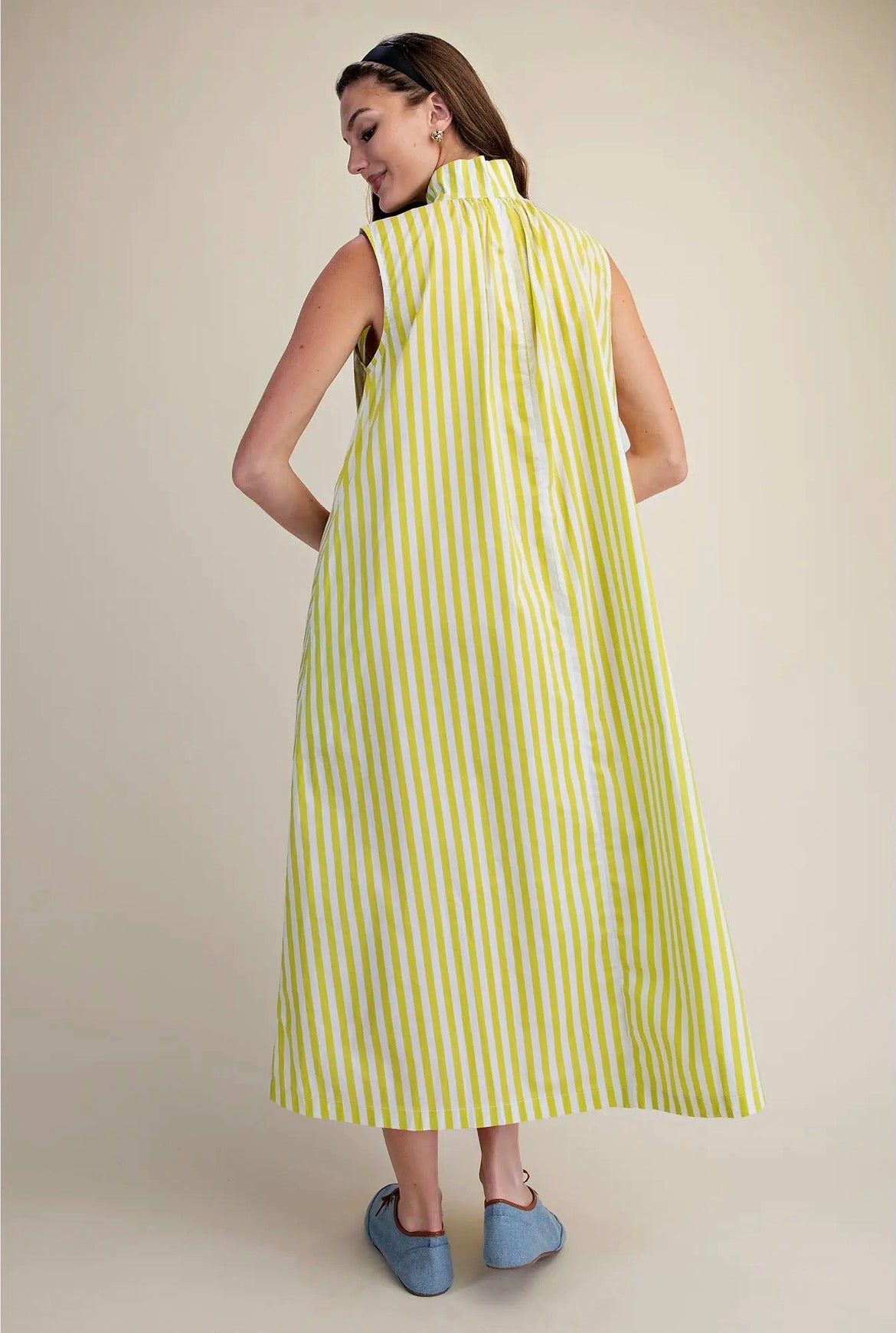 A woman wearing a sleeveless, high-neck midi dress with vertical lime green and white stripes, shown from the back against a solid beige studio background.