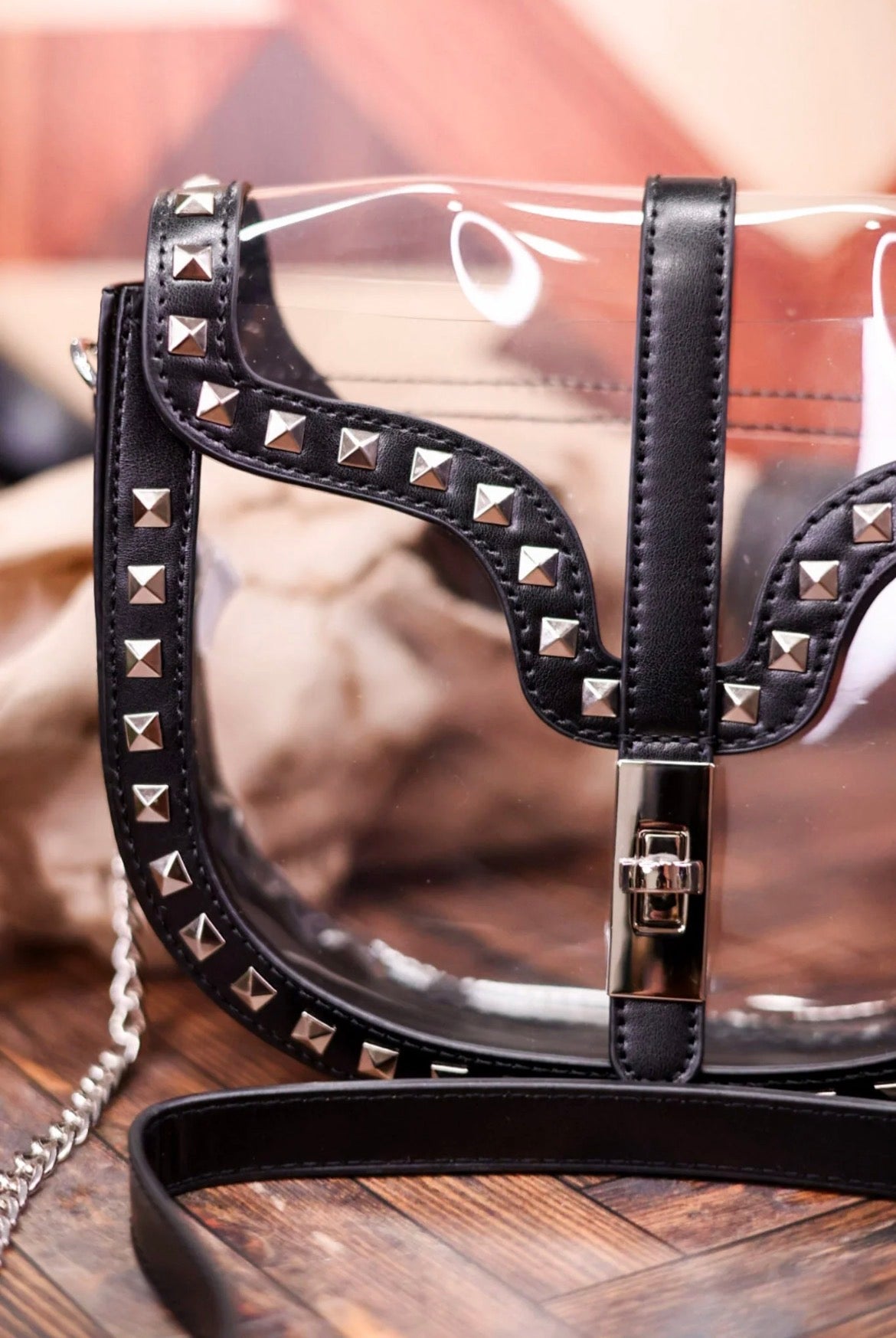 A clear vinyl shoulder bag with black faux-leather trim, silver pyramid studs, and a black strap, shown on a wooden surface.