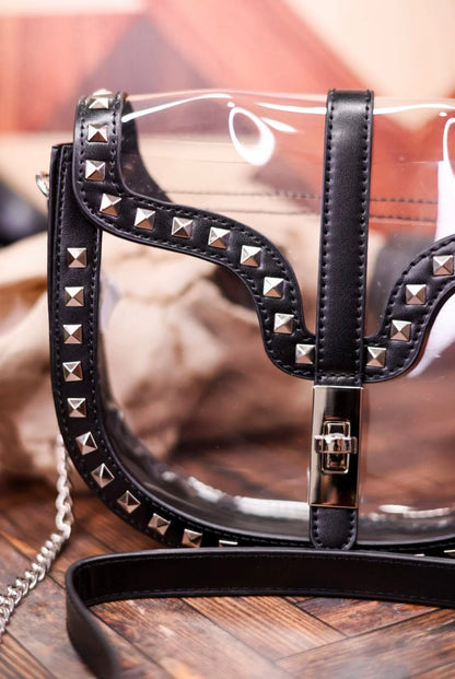 A clear vinyl shoulder bag with black faux-leather trim, silver pyramid studs, and a black strap, shown on a wooden surface.