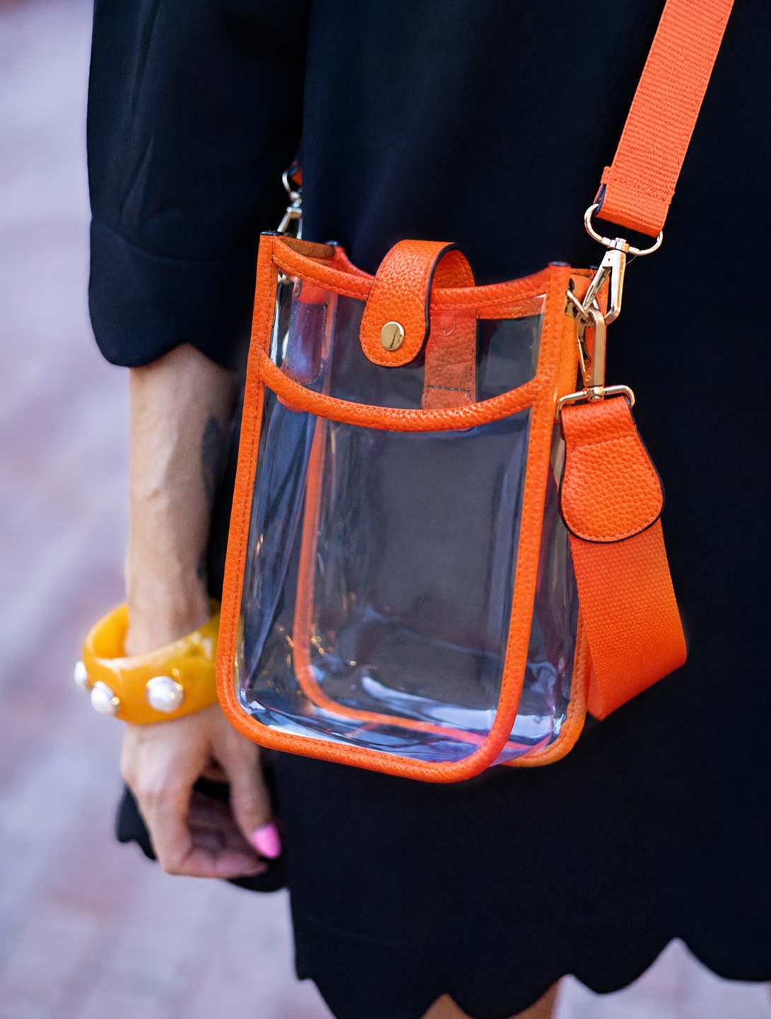 A clear plastic bucket-style shoulder bag with bright orange trim and strap, shown worn crossbody against a person in a black dress. The bag has gold-tone hardware and a snap-button closure.