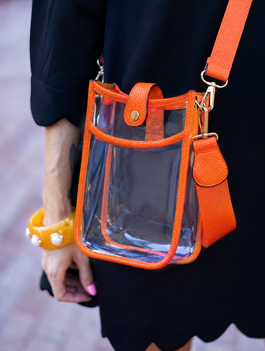 A clear plastic bucket-style shoulder bag with bright orange trim and strap, shown worn crossbody against a person in a black dress. The bag has gold-tone hardware and a snap-button closure.