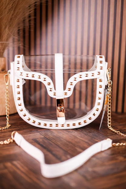 A white faux-leather handbag with a clear acrylic front panel, decorated with gold-tone studs and a gold chain strap, resting on a dark wooden surface.