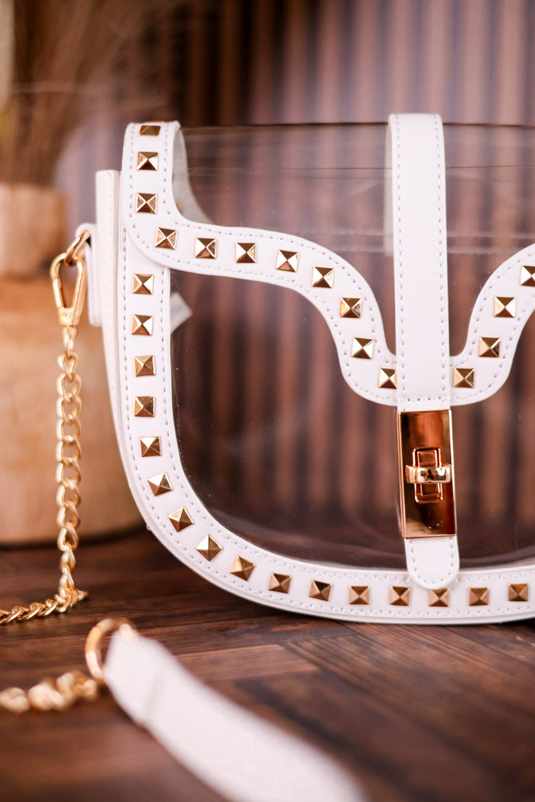 A white faux-leather shoulder bag with gold pyramid studs and a gold chain strap, shown on a dark wooden surface with a blurred background.