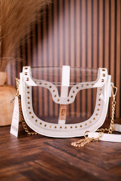 A white faux-leather crescent-shaped shoulder bag with a clear plastic front panel, gold-tone studs, and a gold chain strap, displayed on a wooden surface.