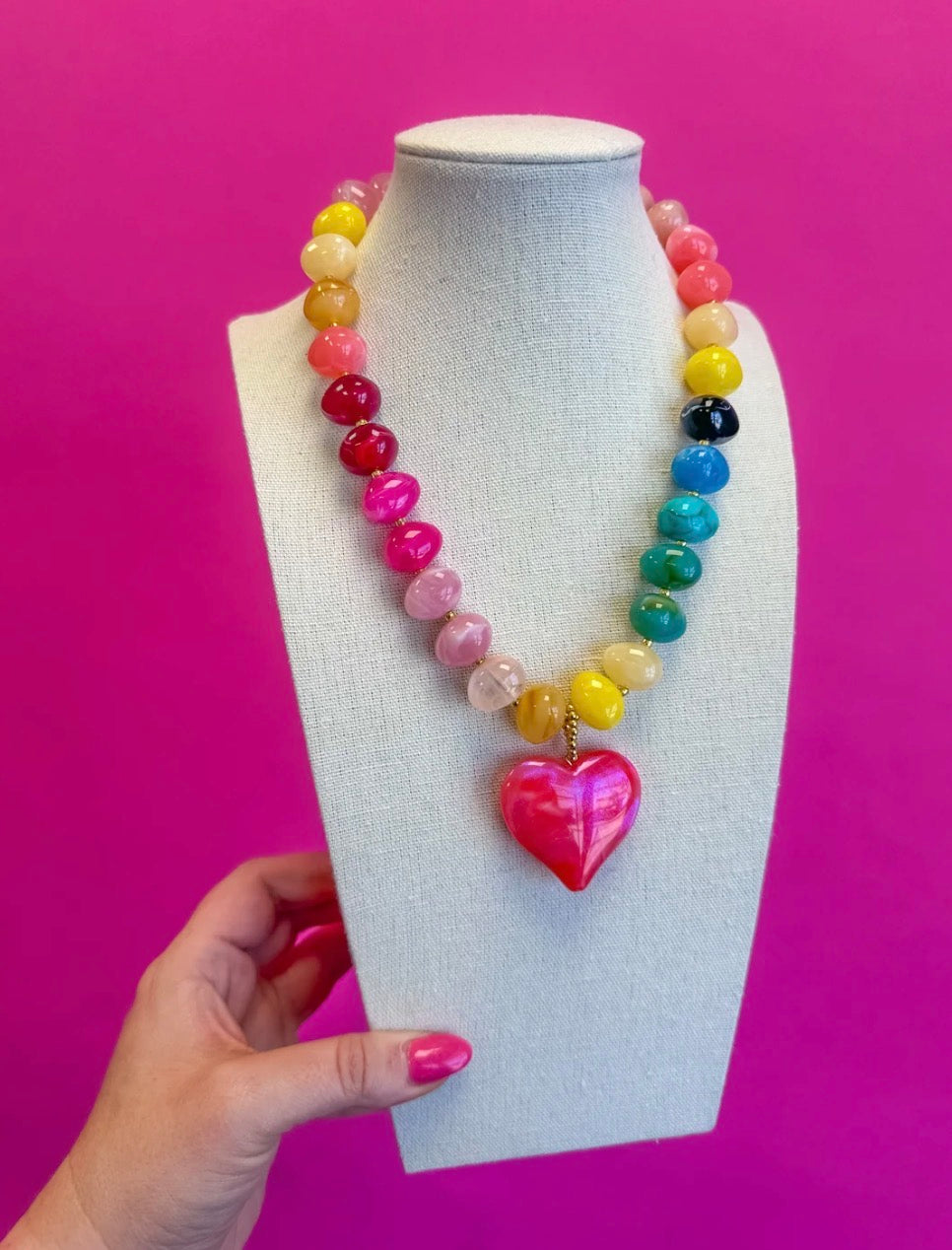 A colorful beaded necklace with a glossy pink heart pendant, displayed on a white fabric mannequin bust. A hand with pink nail polish is holding the base of the bust against a solid bright pink background.