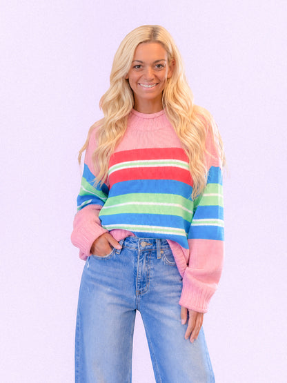 A woman with long blonde hair wearing a pastel striped knit sweater with pink, red, blue, and green horizontal stripes, paired with light-wash blue jeans, against a solid light lavender background.