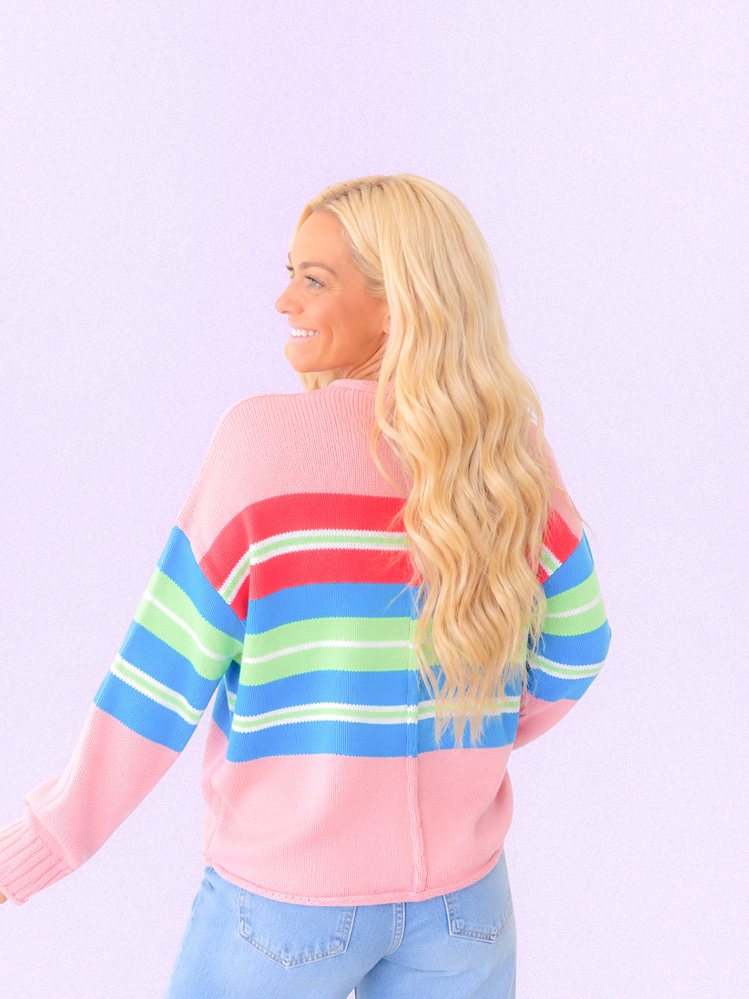 A woman with long blonde hair is shown from the back wearing a pink knit sweater with horizontal stripes in red, blue, and green, paired with light blue jeans, against a solid light purple background.