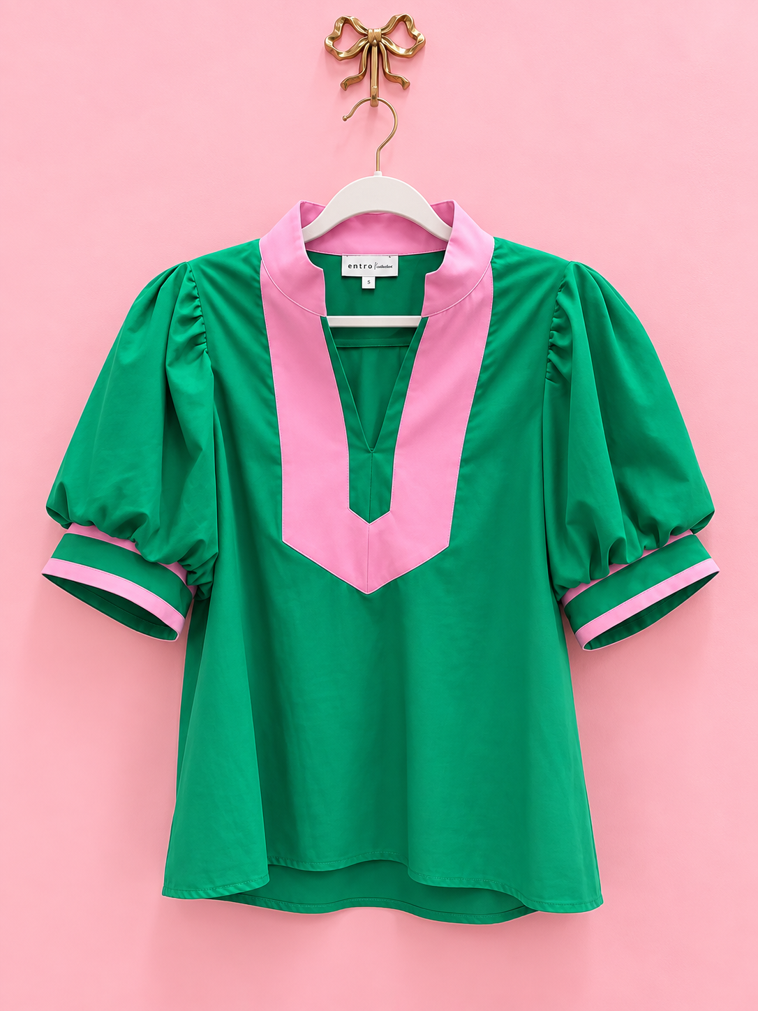 A green short-sleeve blouse with a pink V-neckline and pink striped cuffs, hanging on a white hanger against a solid pink background.