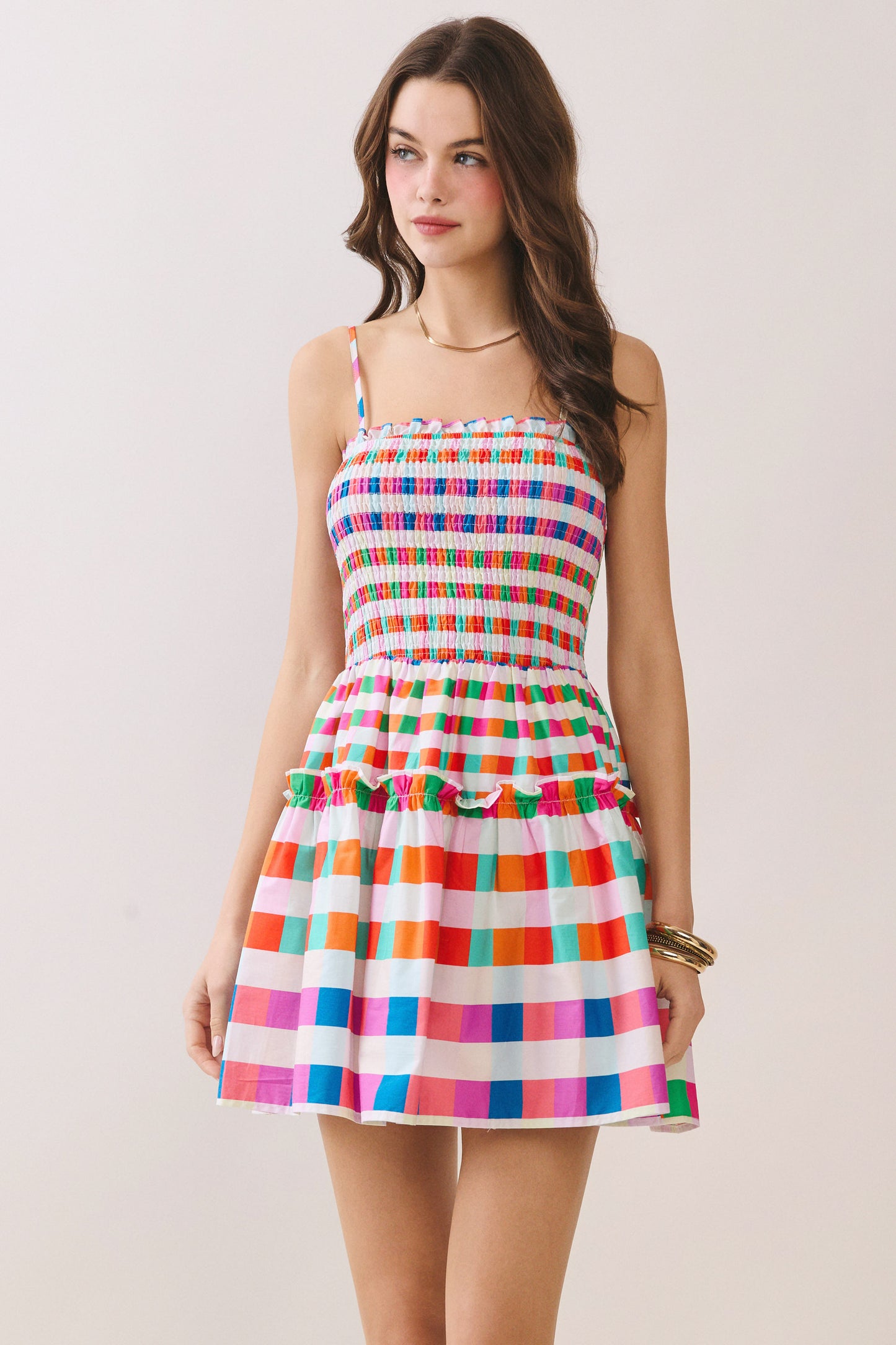 A woman modeling a sleeveless mini dress with thin straps, a smocked bodice, and a tiered skirt in a colorful rainbow stripe pattern on a white background.