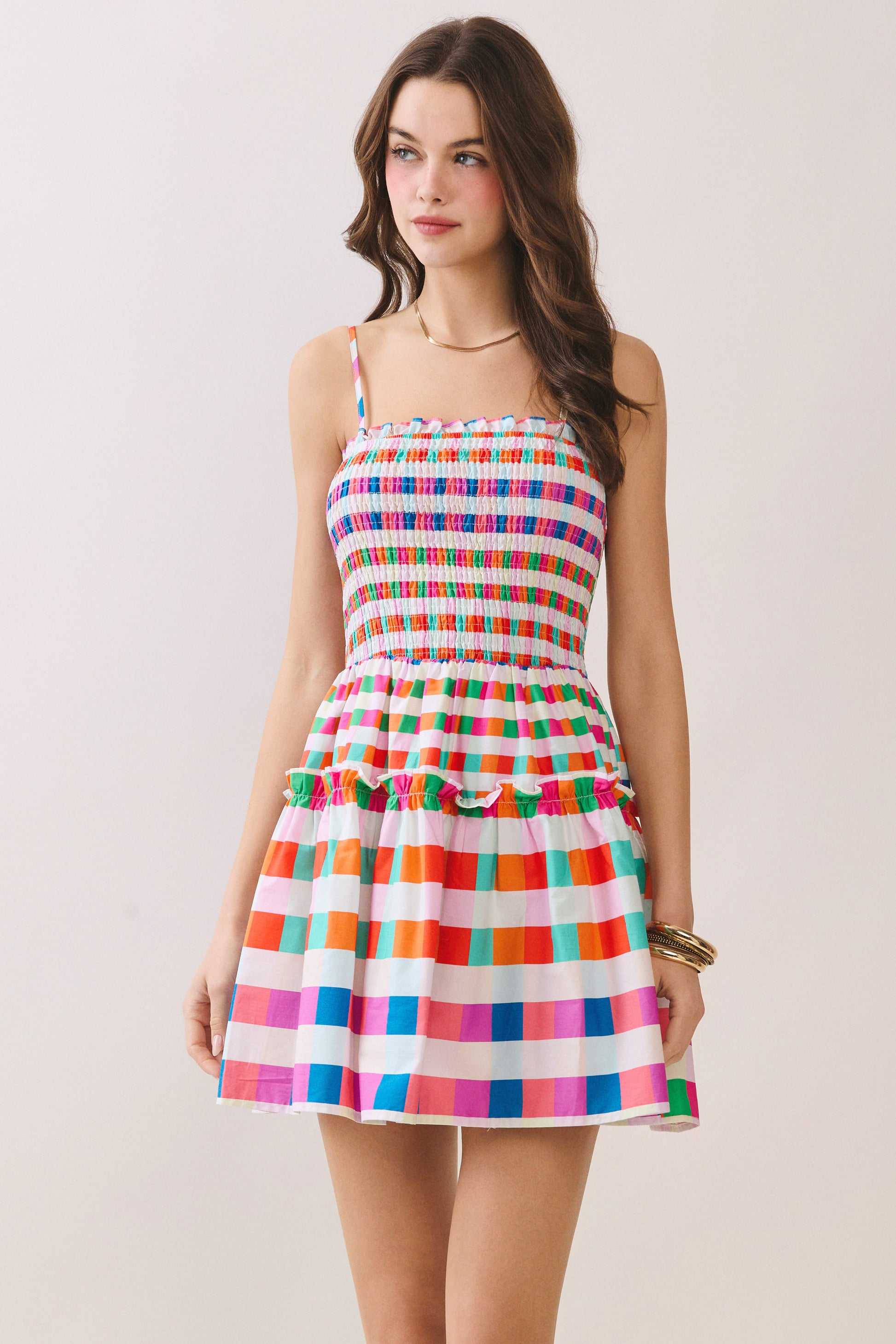 A woman modeling a sleeveless mini dress with thin straps, a smocked bodice, and a tiered skirt in a colorful rainbow stripe pattern on a white background.