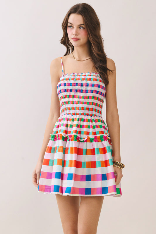 A woman modeling a sleeveless mini dress with thin straps, a smocked bodice, and a tiered skirt in a colorful rainbow stripe pattern on a white background.