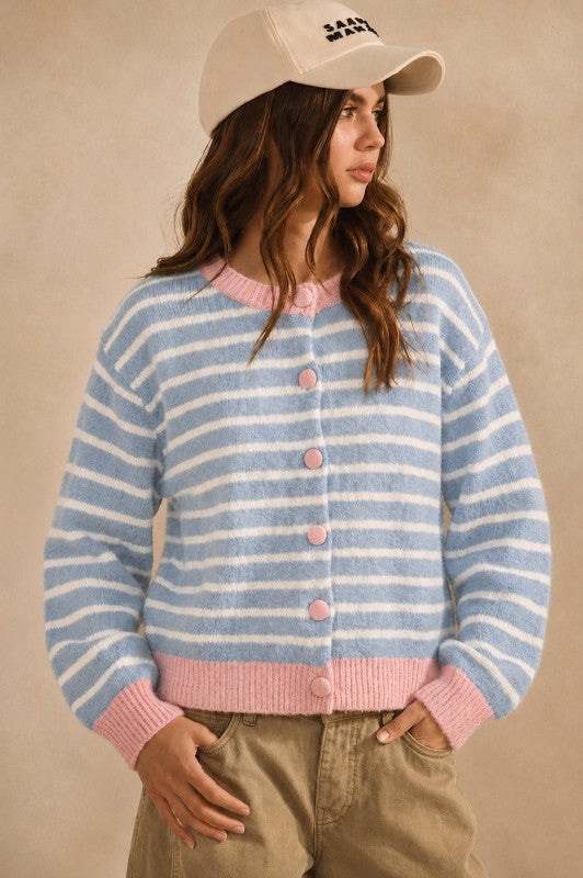 A woman wearing a light blue and white striped fuzzy cardigan with pink trim and buttons, paired with khaki pants and a beige baseball cap, set against a warm beige studio background.