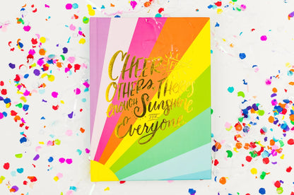 Rainbow striped greeting card with gold text, surrounded by colorful confetti on white background.