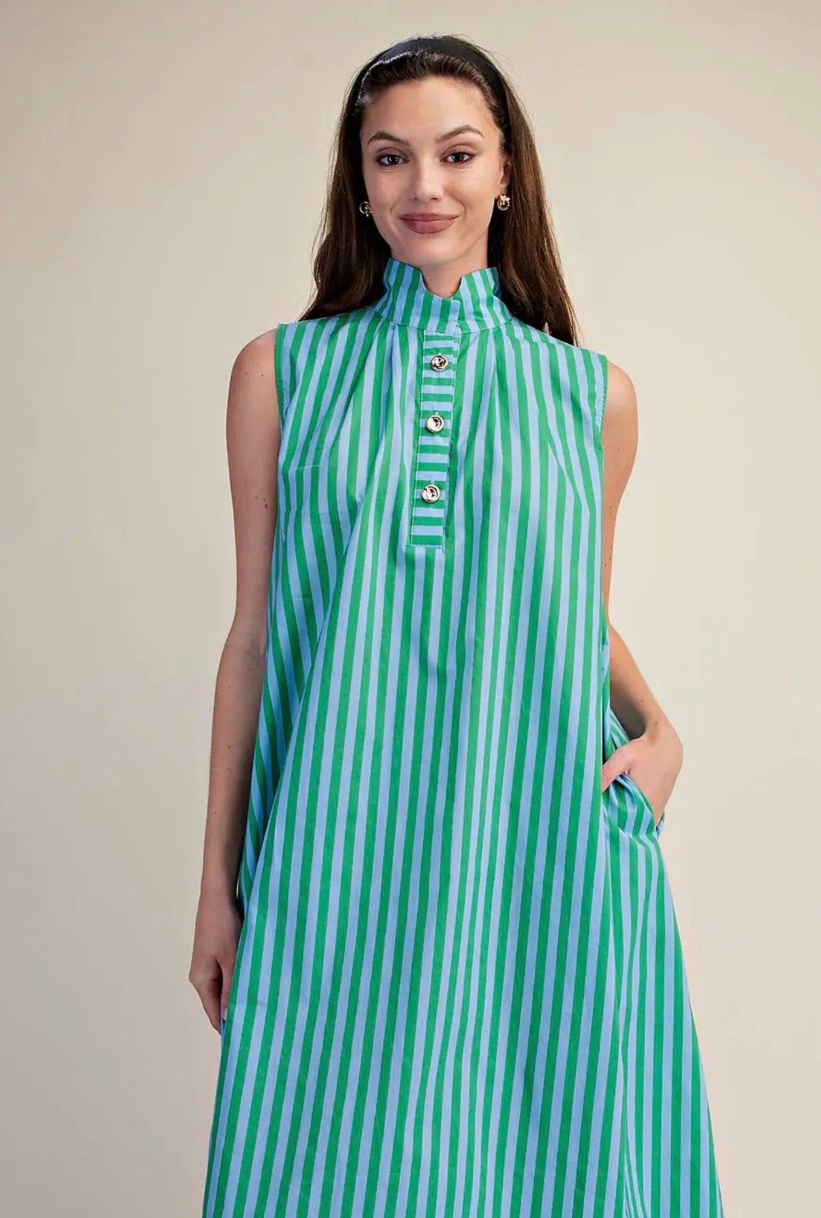 A woman modeling a sleeveless high-neck dress with vertical mint green and white stripes, featuring decorative gold-tone buttons down the front placket, set against a plain beige studio background.