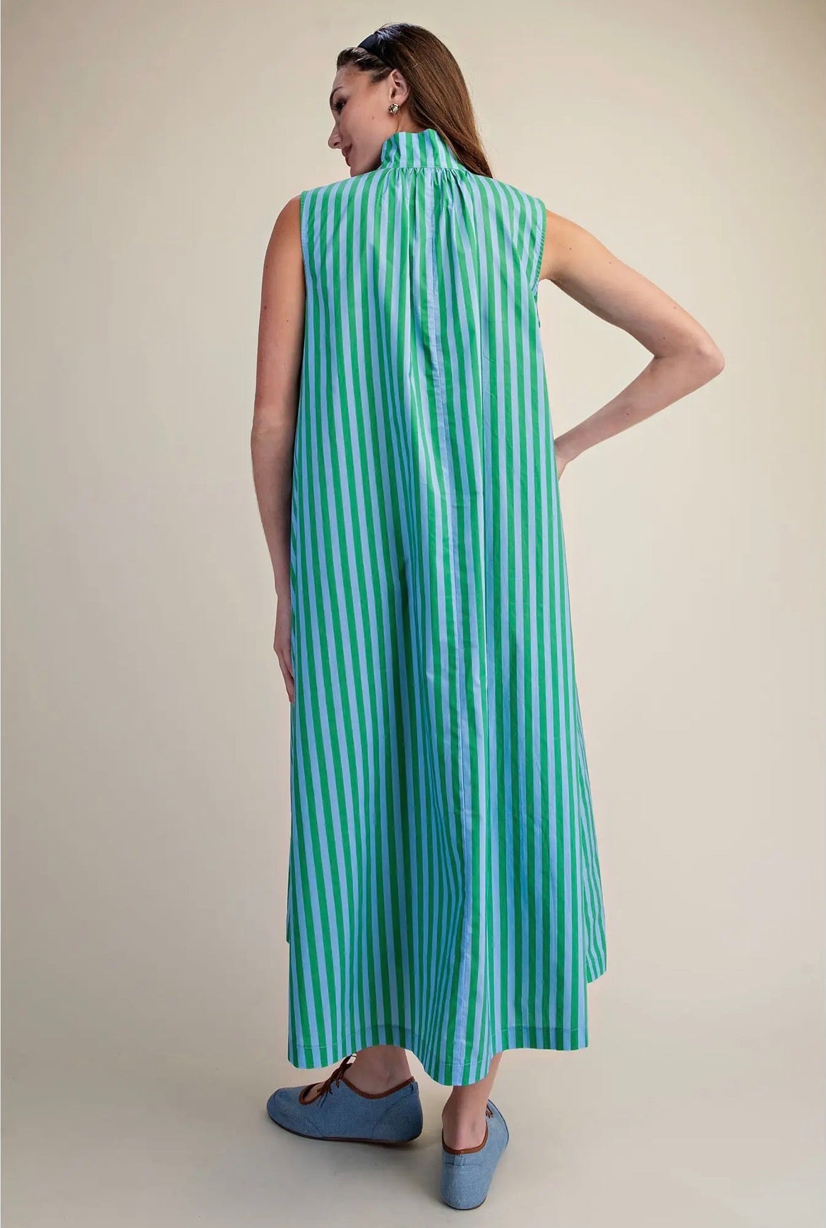 A woman shown from the back wearing a sleeveless high-neck maxi dress with vertical mint green and white stripes, paired with blue slip-on shoes, against a plain beige studio background.