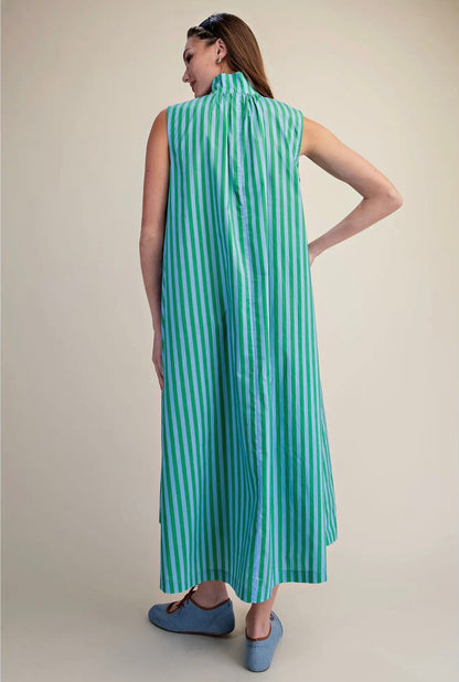 A woman shown from the back wearing a sleeveless high-neck maxi dress with vertical mint green and white stripes, paired with blue slip-on shoes, against a plain beige studio background.