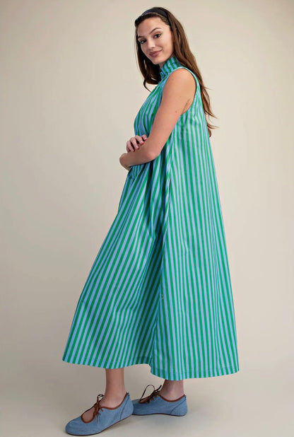 A woman modeling a sleeveless, high-neck midi dress with vertical mint green and white stripes, paired with light blue Mary Jane shoes, against a solid beige studio background.
