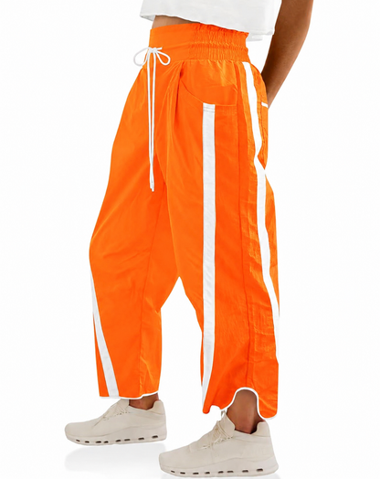 Bright orange wide-leg track pants with white side stripes and a white drawstring waist, shown on a model from the waist down wearing off-white sneakers against a white background.