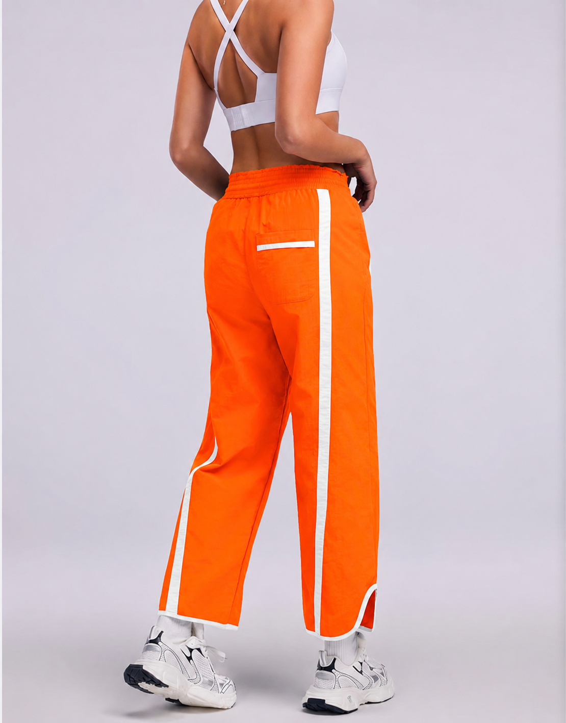 A person shown from the back wearing bright orange cropped pants with white side stripes, a white sports bra, and white sneakers against a light gray studio background.