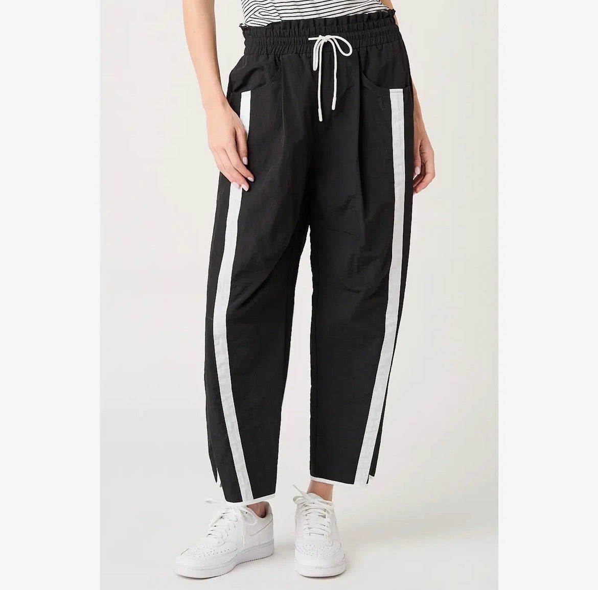 Black cropped pants with white side stripes, worn by a model with white sneakers against a light studio background.